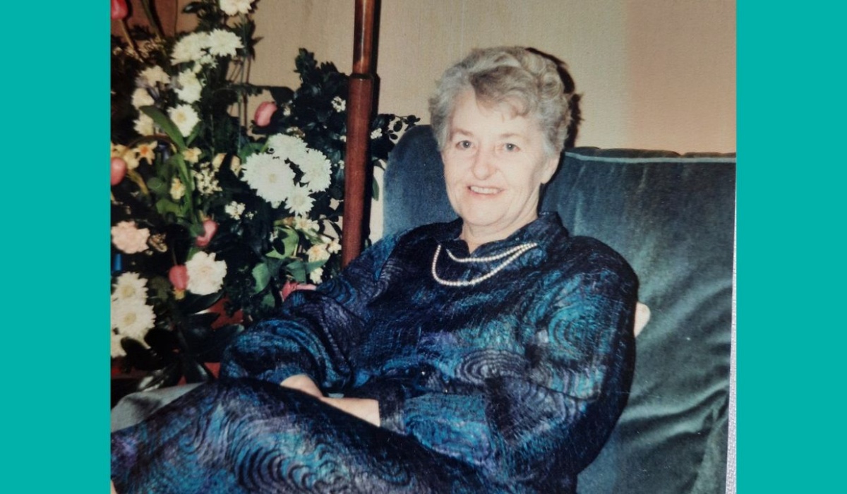 Daphne is dressed in a blue dress and pearls, smiling at the camera, seated on a blue velvet sofa with a large bouquet of flowers next to it.